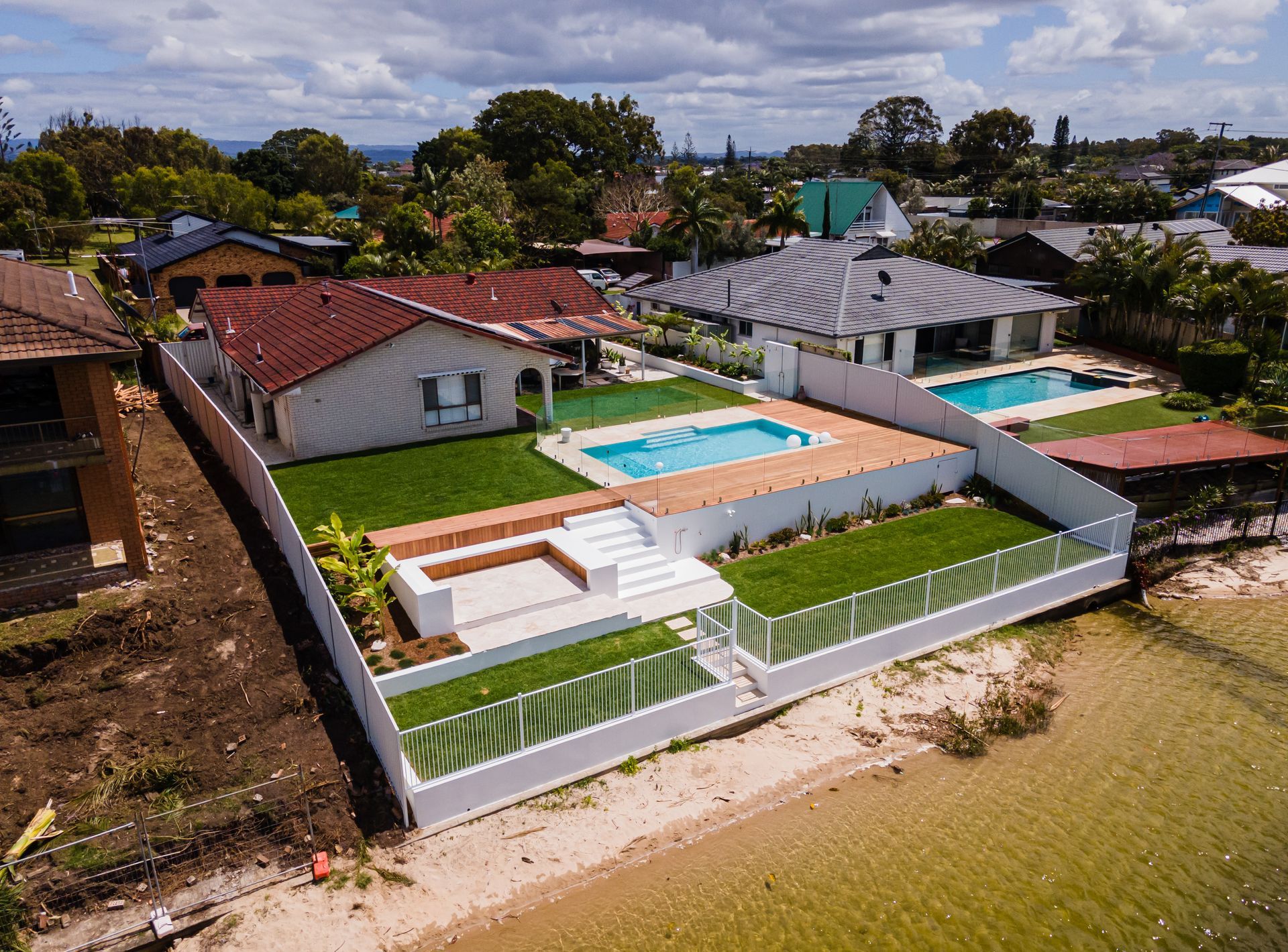 Aerial view of a coastal house with a multi-level backyard, swimming pool, green lawn, and a white fence by the water.