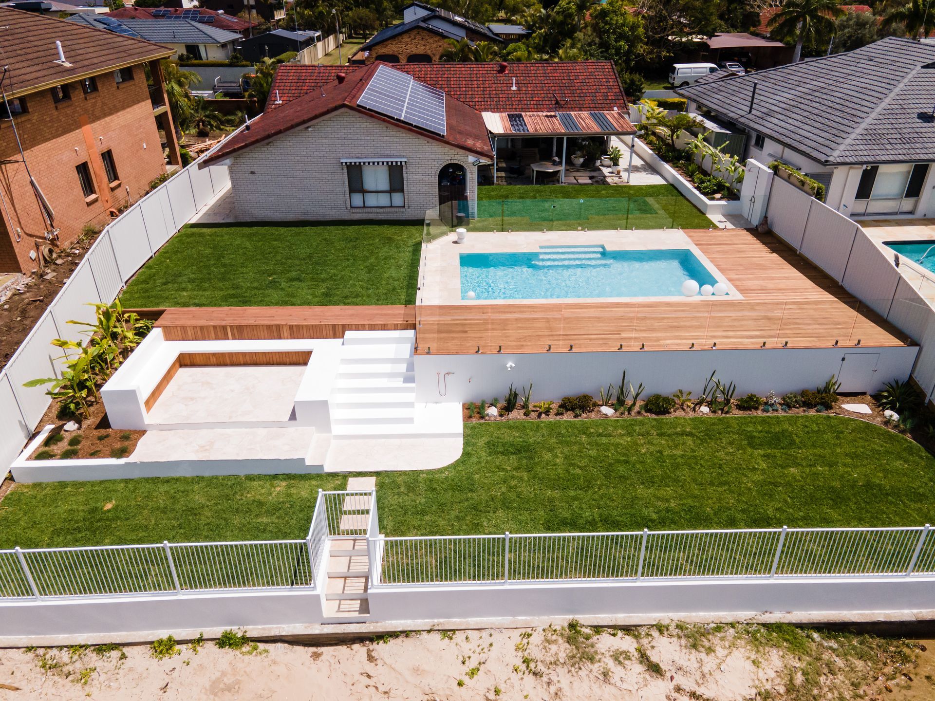 Aerial view of a residential backyard with a swimming pool, wood decking, white retaining walls, and a lawn.
