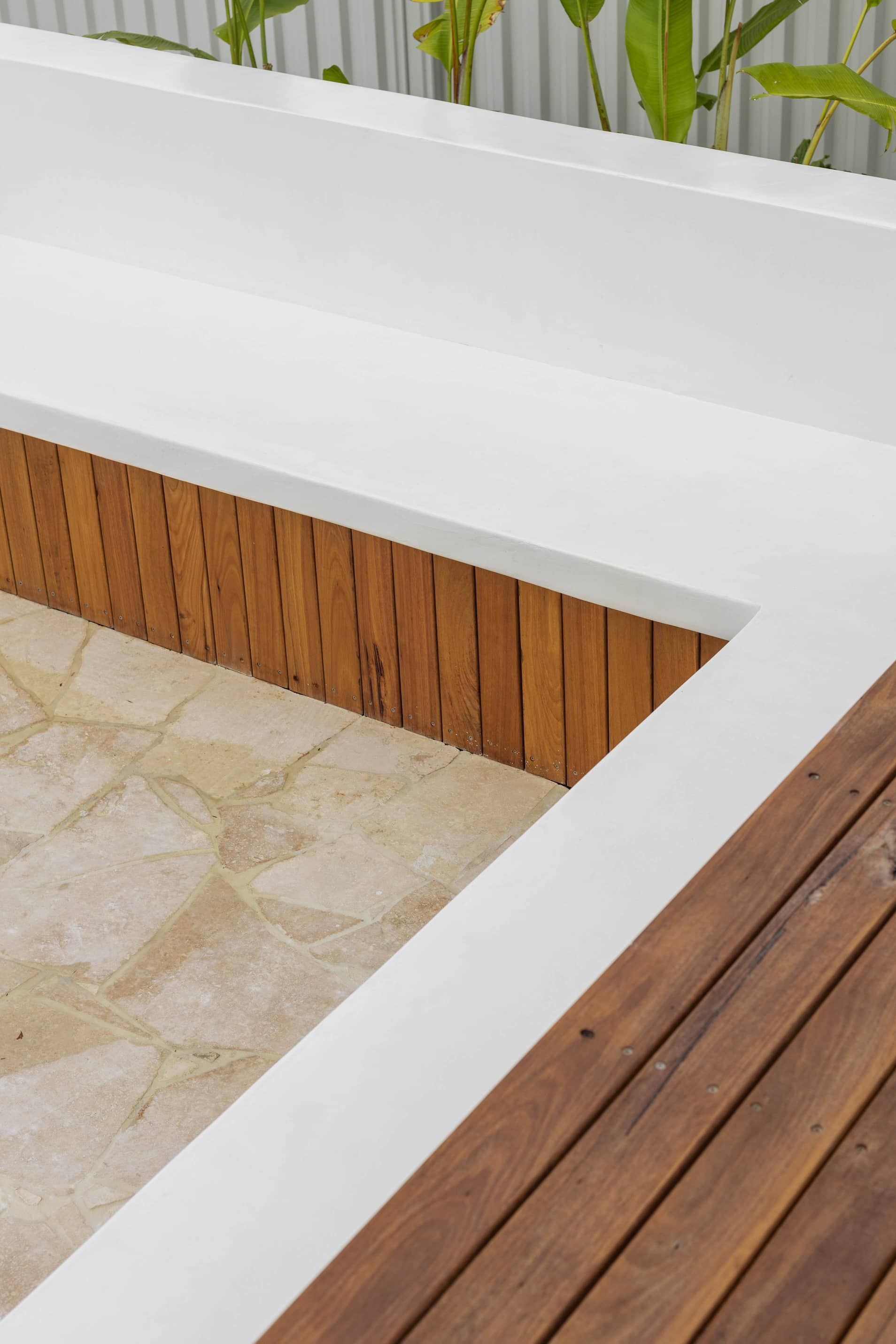 A white L-shaped concrete bench with a slatted wood base sits next to a wooden deck and a light-colored stone patio.