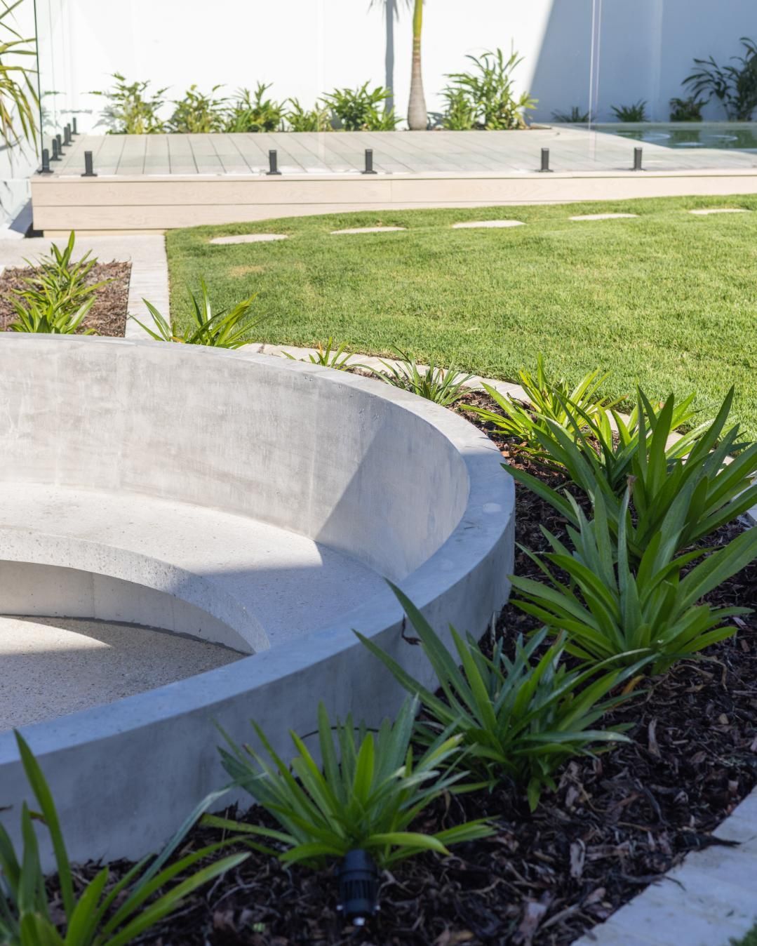 A curved concrete bench sits in a garden bed with spiky plants and mulch, overlooking a grassy lawn and wood deck.