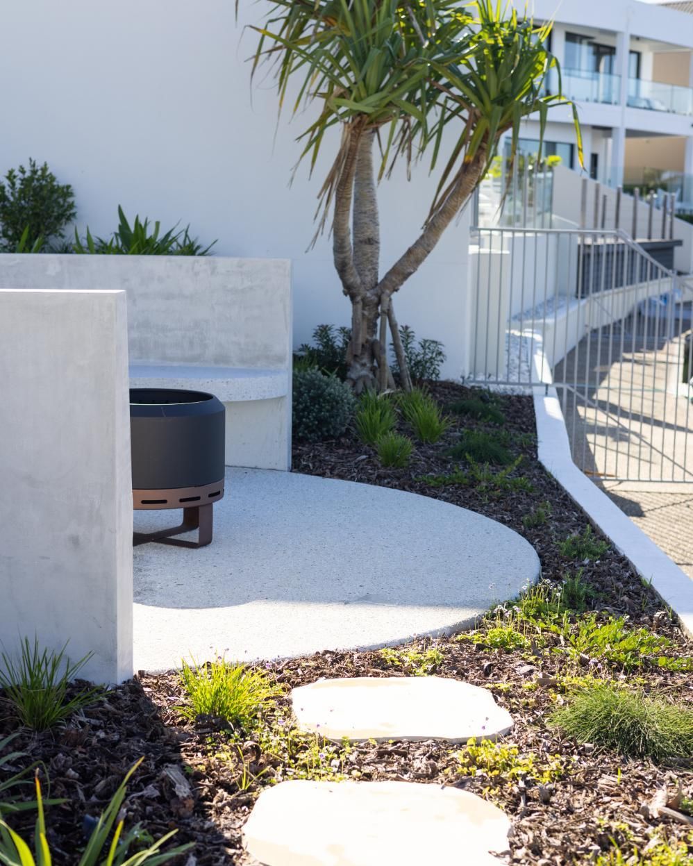 A modern patio with a white curved bench, a dark fire pit, and stone stepping stones set in a landscaped yard.