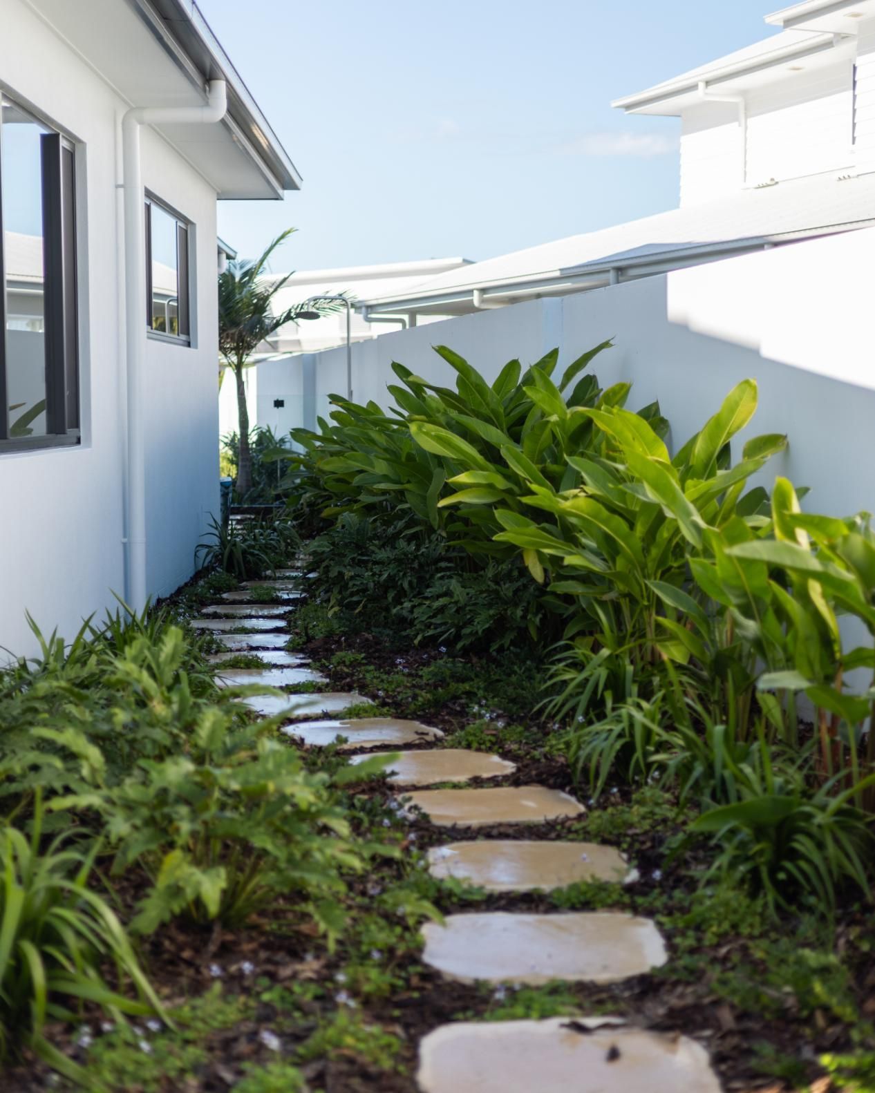 A stepping stone path leads through a narrow, landscaped garden between a white house and a tall white wall.