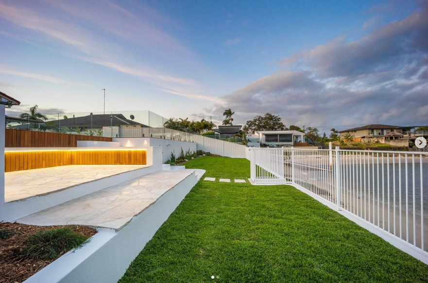 A tiered outdoor patio with wooden bench lighting, green grass, and a white fence overlooking a canal at sunset.