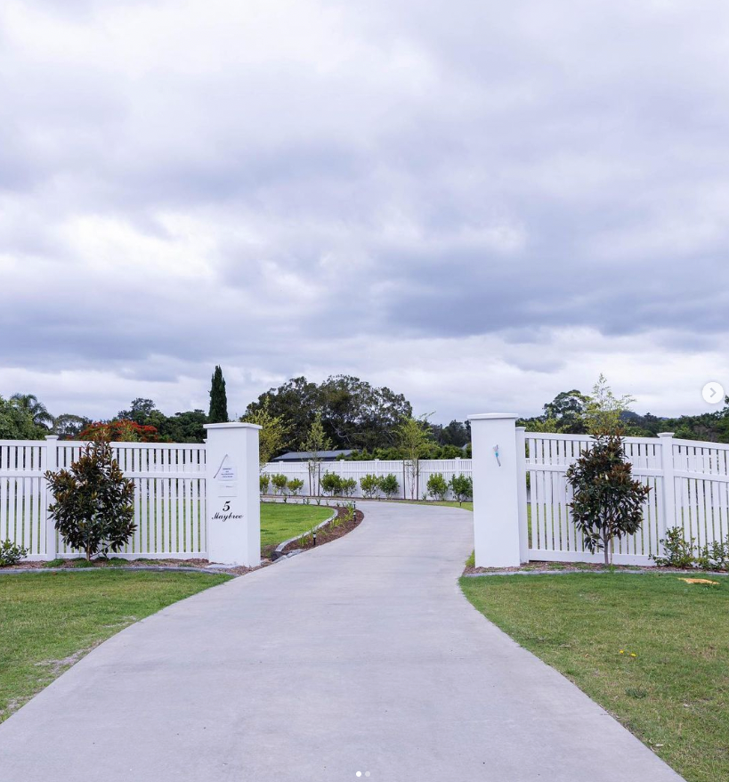 A paved driveway leads through a white picket fence gate flanked by two white pillars on a cloudy day.