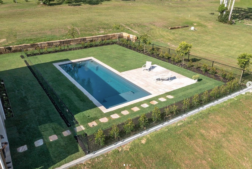 An aerial view of a rectangular blue swimming pool surrounded by green turf, a light-colored patio, and a black fence.