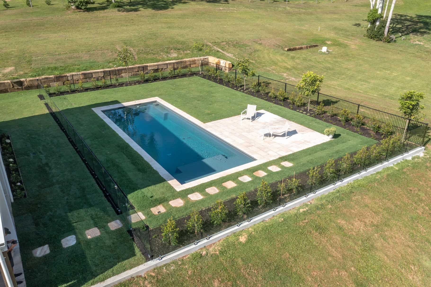 An aerial view of a rectangular backyard pool with a lounge area, surrounded by a green lawn and a perimeter fence.
