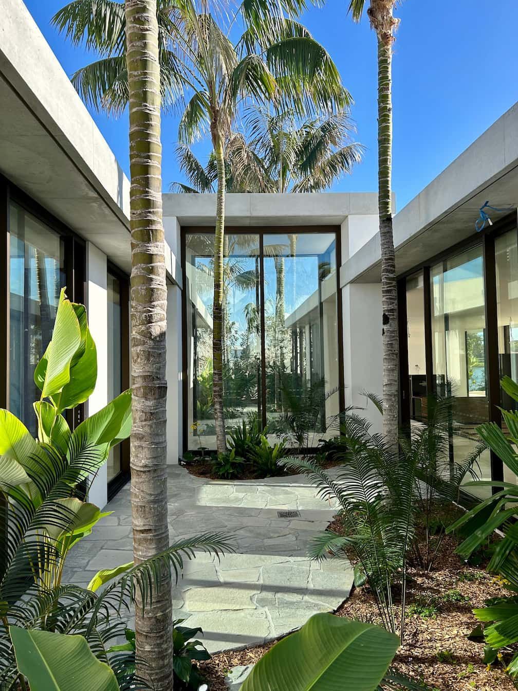 A modern home with white concrete walls and floor-to-ceiling glass windows surrounding a courtyard garden with palm trees.