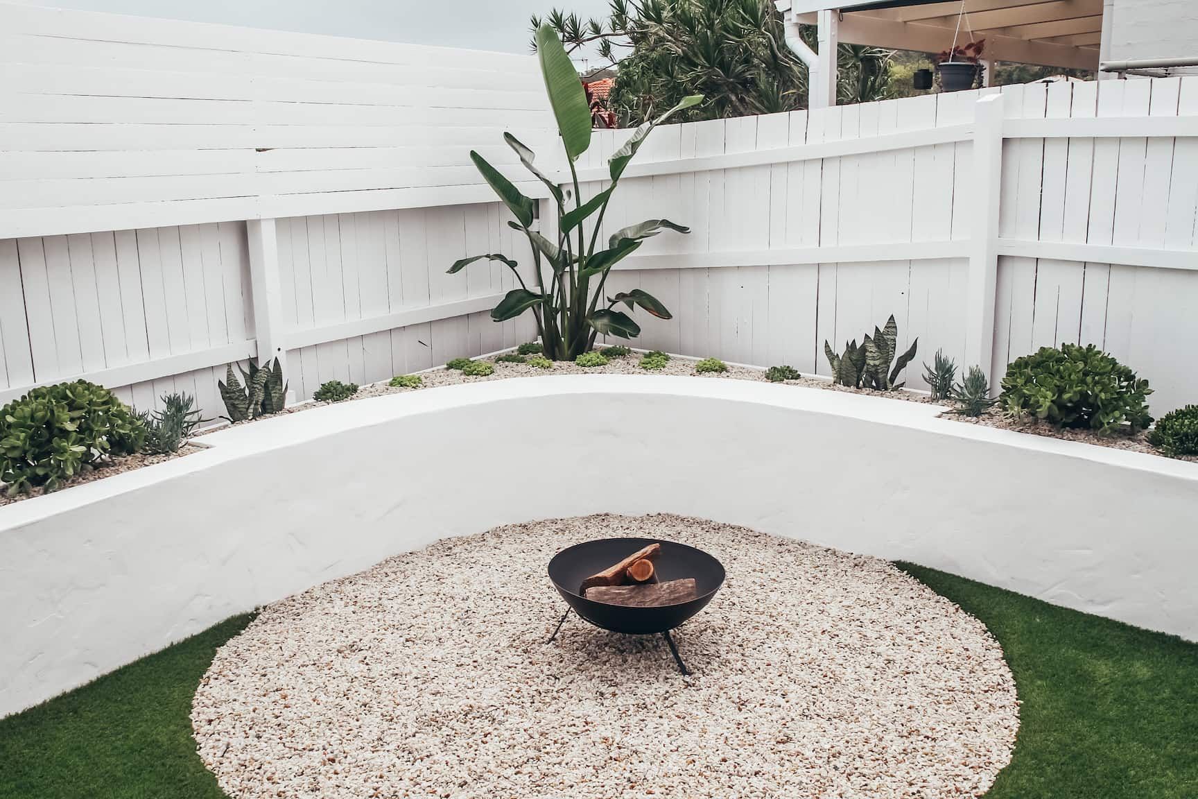 A fire pit centered on a circular gravel area, surrounded by a curved white wall, green plants, and a white privacy fence.