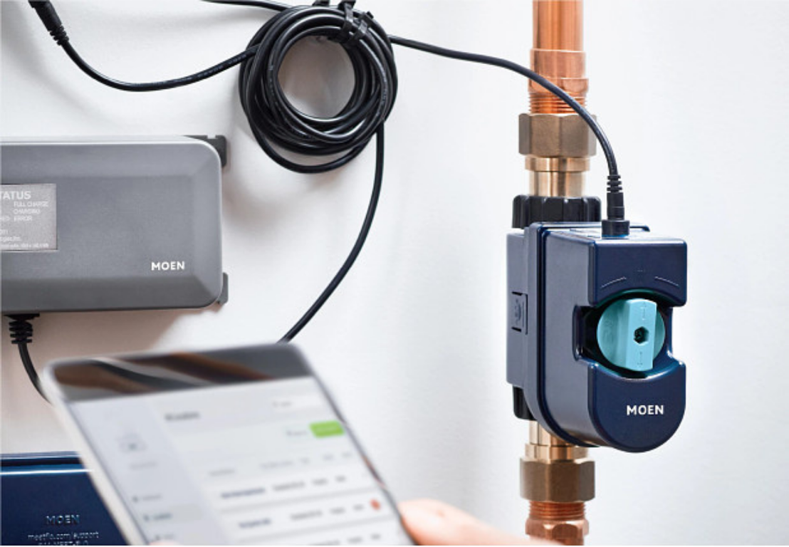 A person is holding a cell phone next to a smart water shutoff. Flow By Moen by DrainWorks Plumbing.