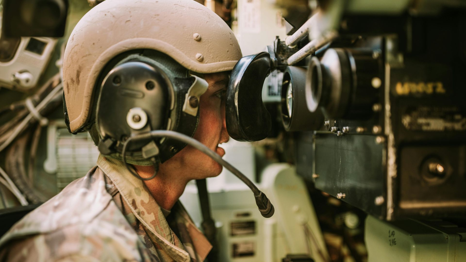 Soldier in helmet and night vision goggles inside a military vehicle, looking forward.