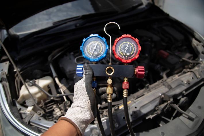 Gloved hand holding AC gauge manifold in front of a car engine bay, checking system pressures.