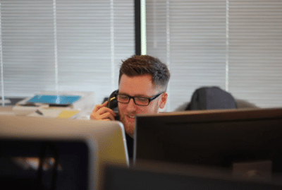 A man wearing glasses is talking on a cell phone in front of a computer.