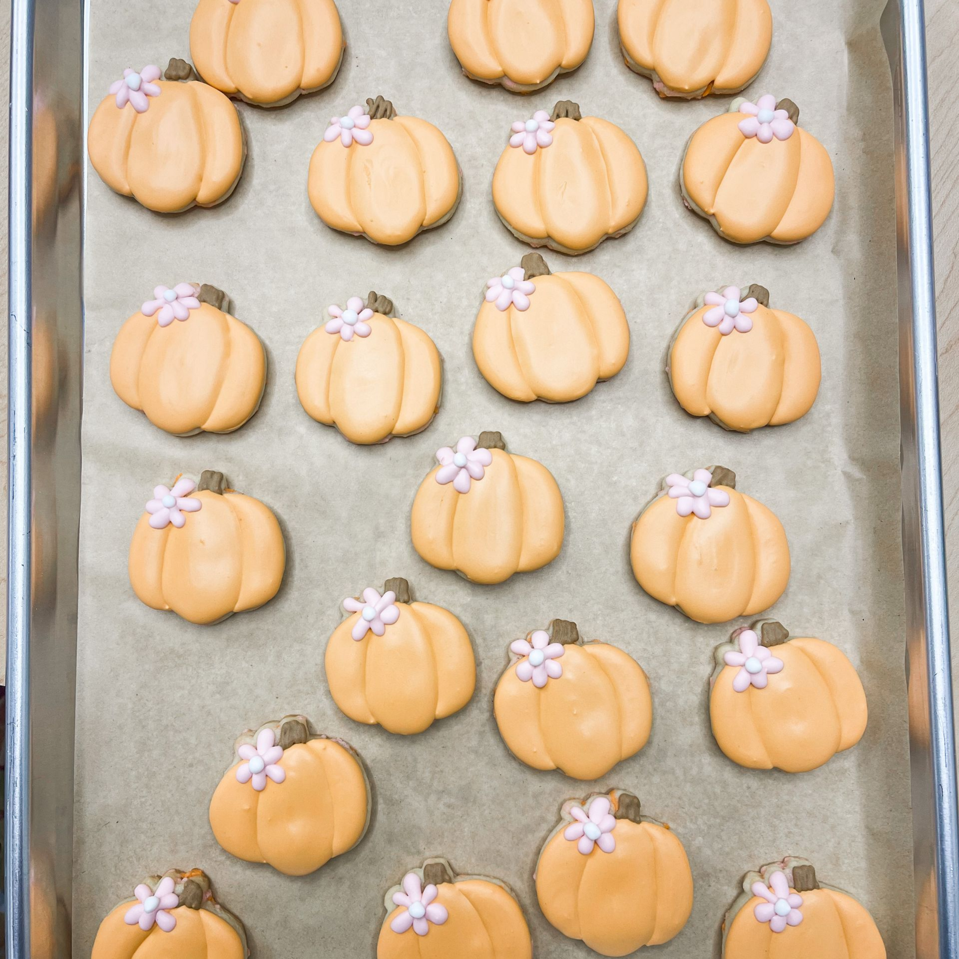 A baking sheet lined with parchment paper holds twenty orange pumpkin-shaped sugar cookies, each decorated with a flower.