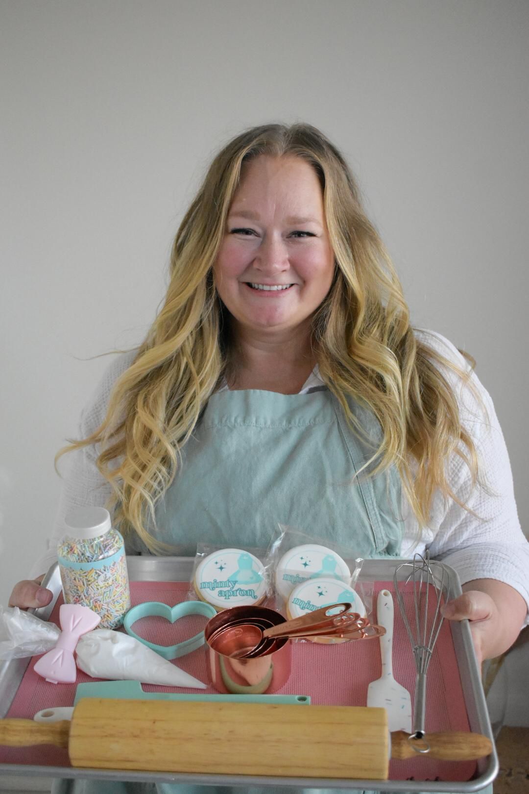 Woman smiling, holding baking sheet with decorated cookies and tools.