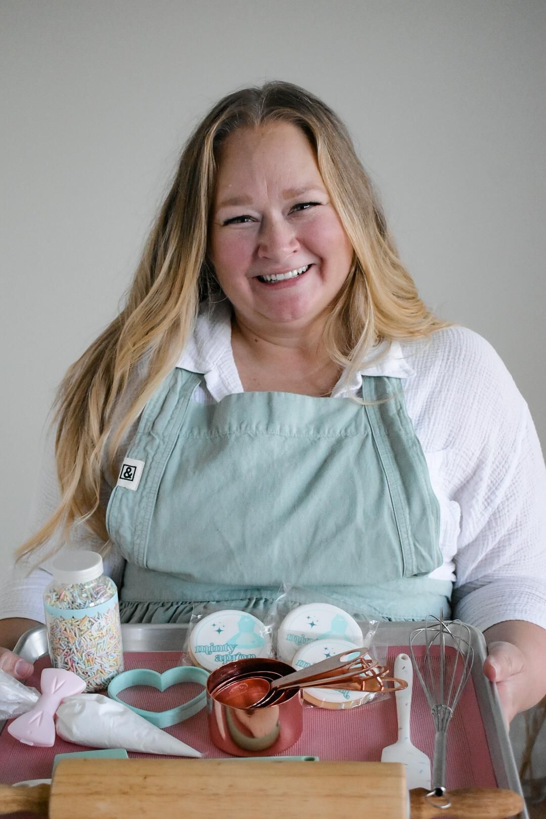 Woman smiling, holding a tray with decorated cookies and baking supplies; light blue apron.