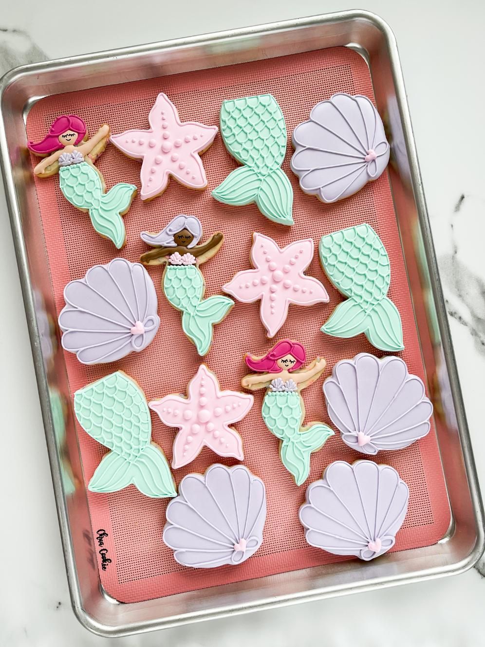 Mermaid-themed cookies: mermaid, starfish, shell shapes in pastel colors on a pink mat in a pan.