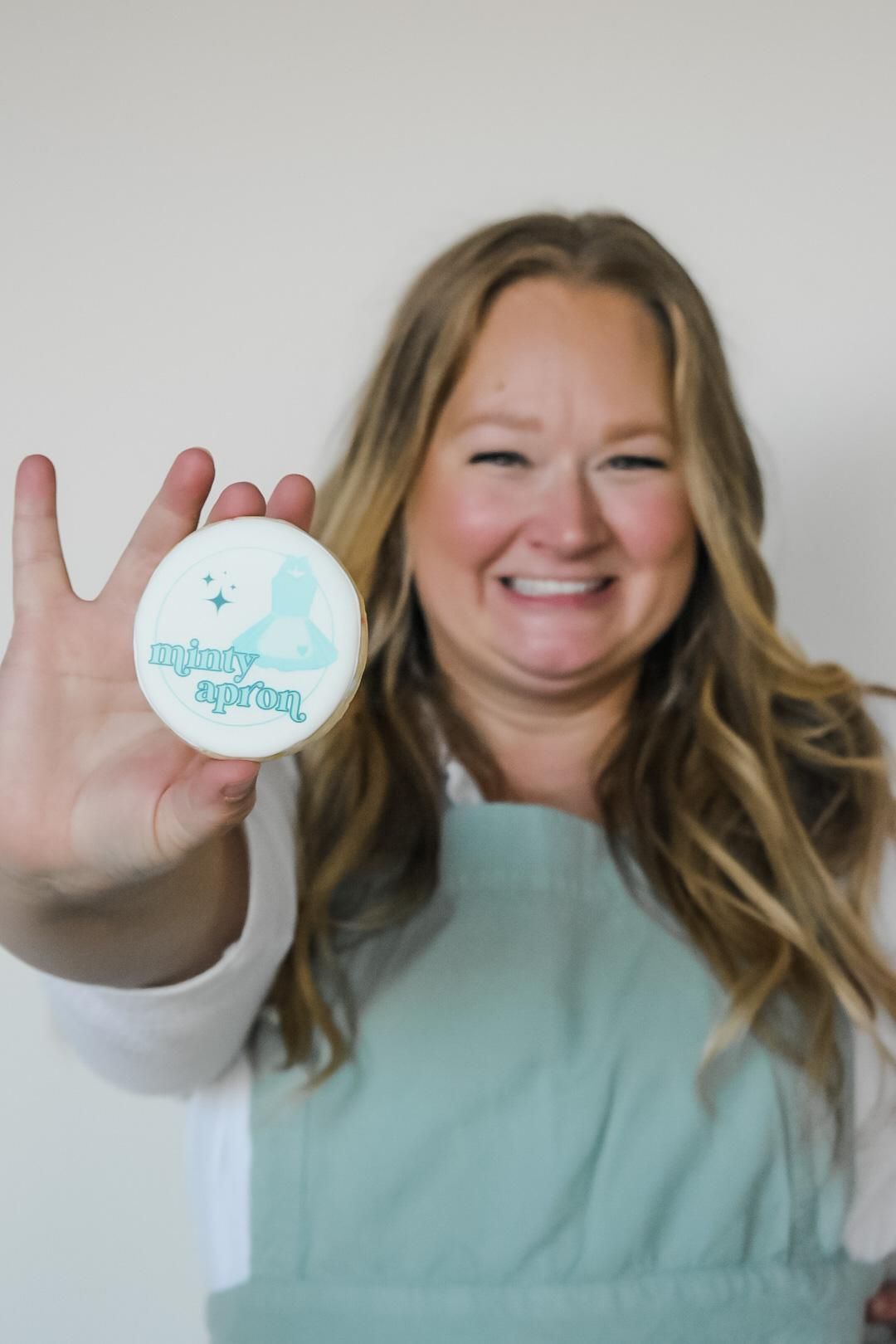 Woman smiling, holding decorated cookie; cookie design includes text, light blue colors.