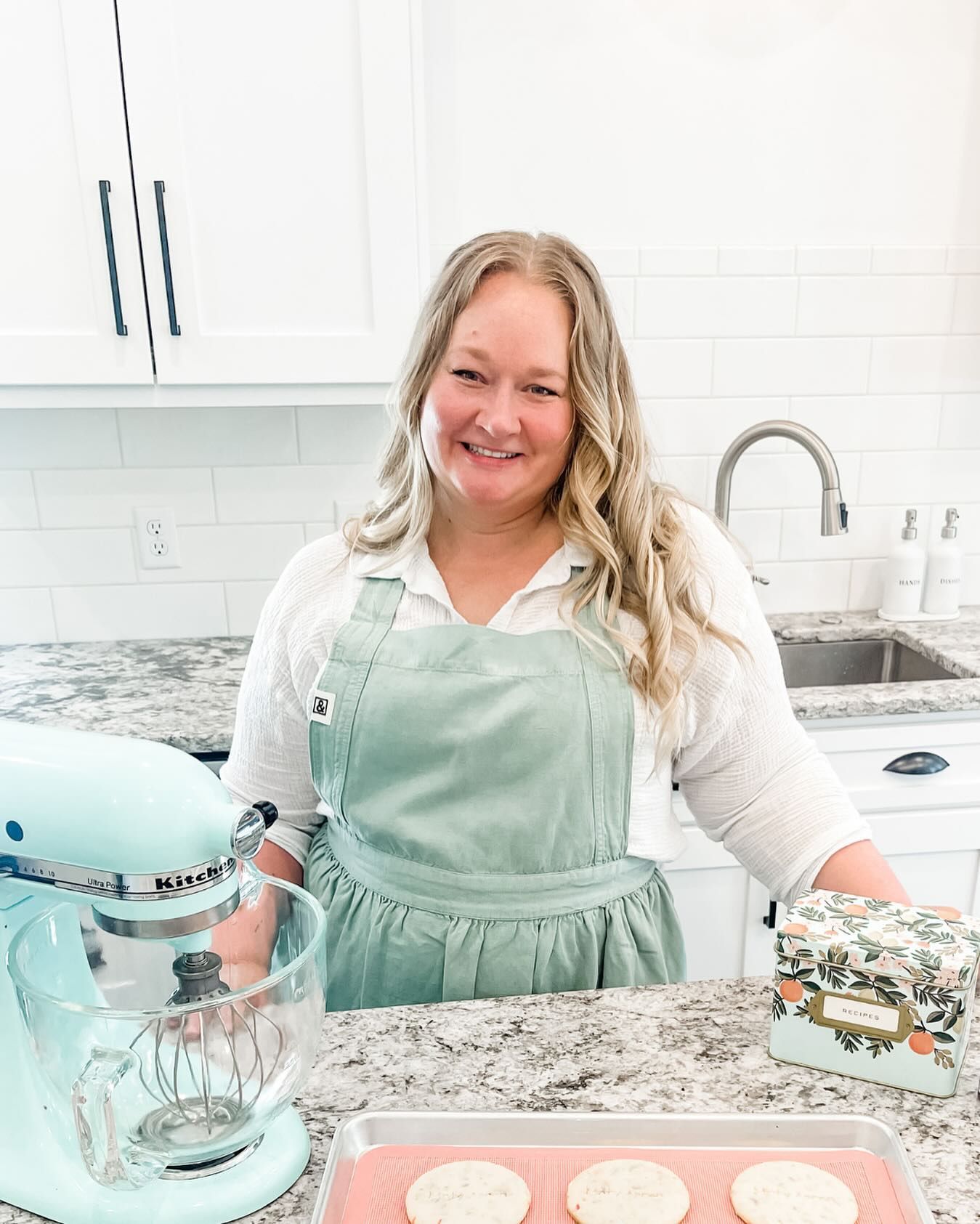 Woman in apron smiles in kitchen, next to mixer and cookies on a tray.
