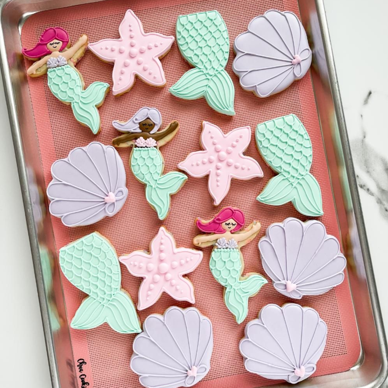 A baking sheet filled with decorated mermaid-themed sugar cookies: mermaids, starfish, seashells, and fishtails.