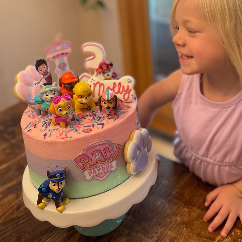 A smiling child looks at a multi-colored Paw Patrol themed birthday cake featuring plastic figures and a name topper.