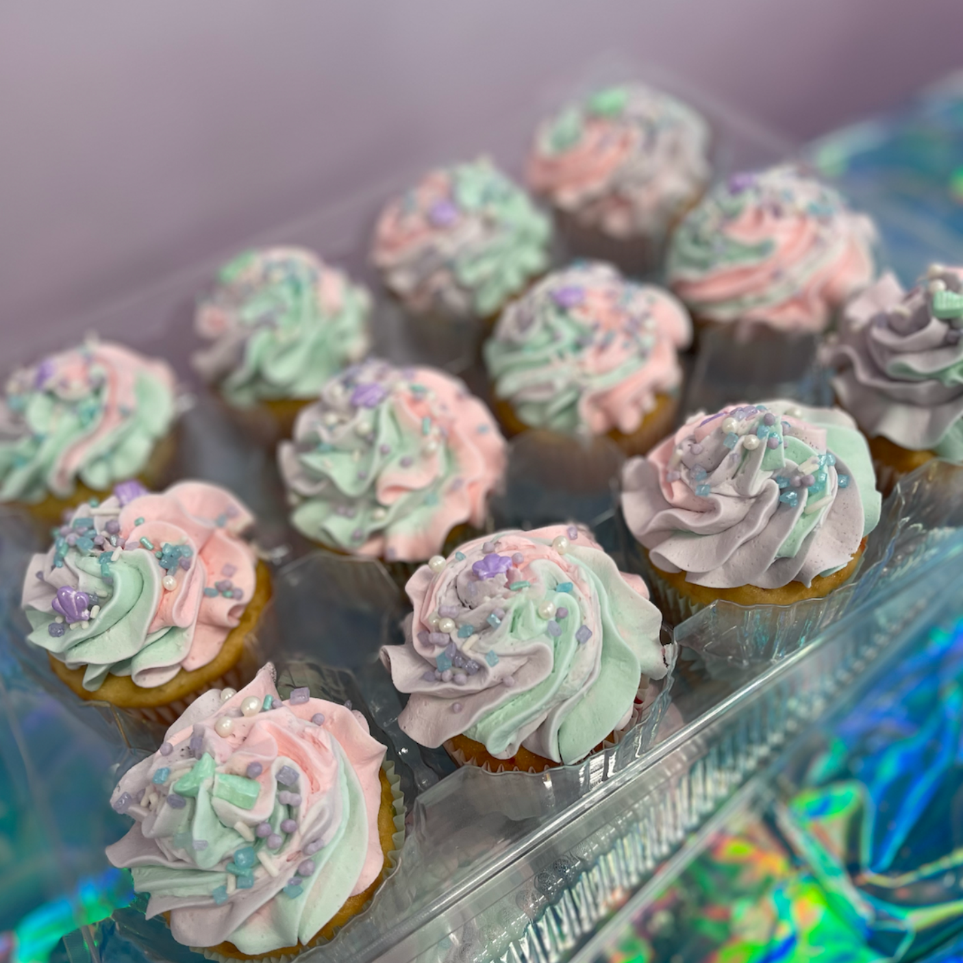 A dozen cupcakes with pastel pink, mint, and lavender swirl frosting and sprinkles, displayed in a clear plastic container.