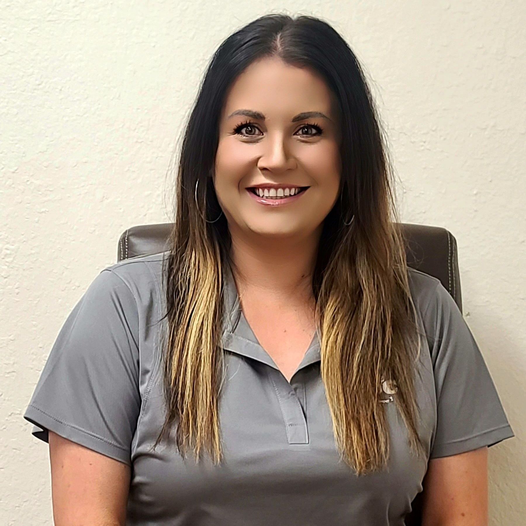 A woman in a grey shirt is smiling while sitting in a chair.