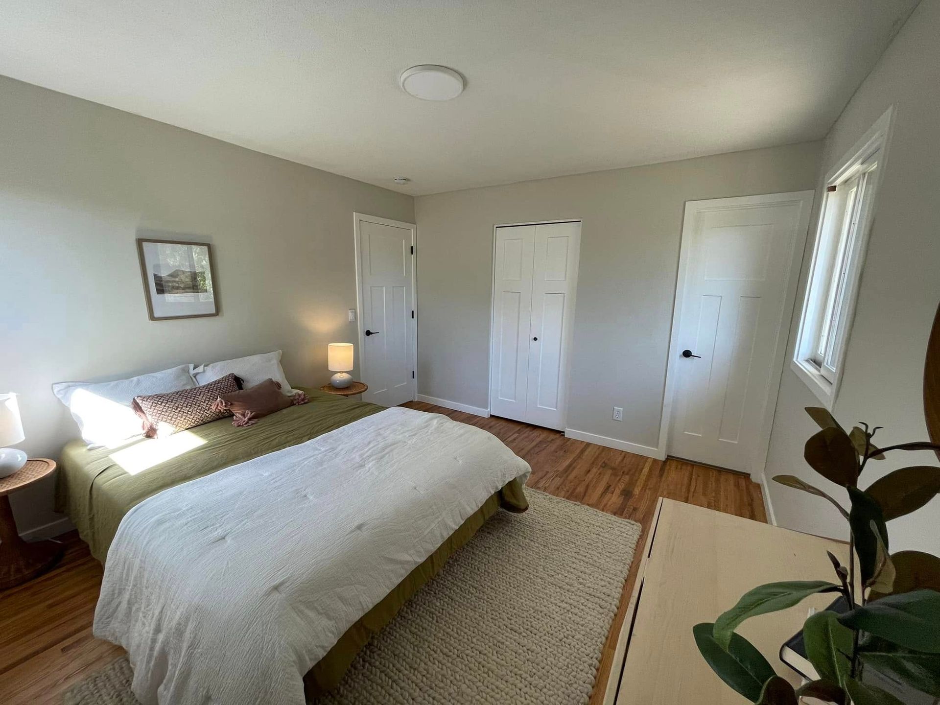 Bedroom with a bed, two closets, two doors, and a window. Beige walls and wooden floor.