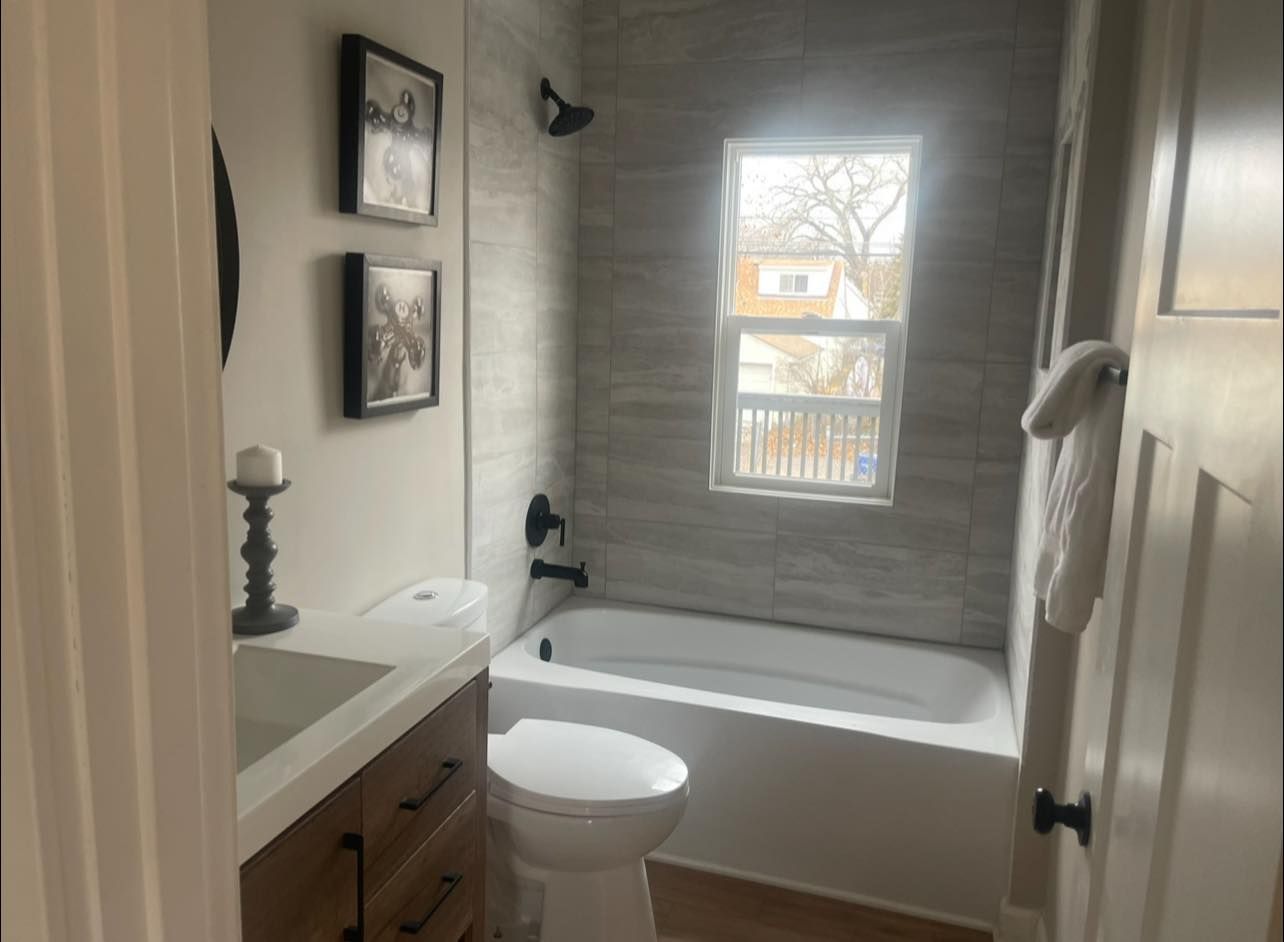 Bathroom with gray tile, white tub, toilet, and vanity. Black fixtures, art, and a small window.