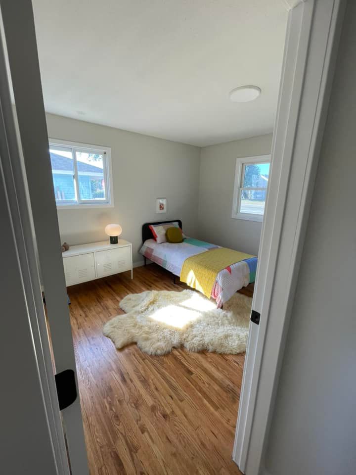 Bedroom with hardwood floors, bed, and white rug; light gray walls, white trim and two windows.
