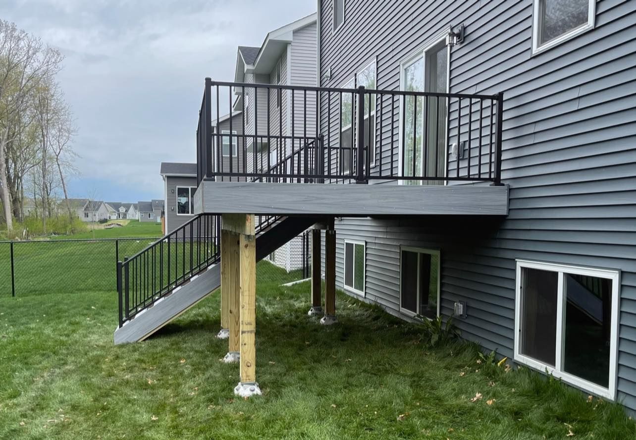 Gray and black deck attached to a gray house, supported by wooden posts, with grass.