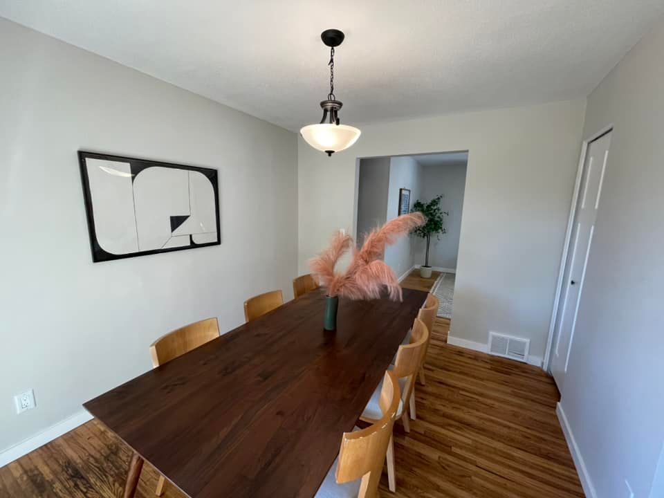 Dining room with long wooden table, light wood chairs, artwork, and pink pampas grass.