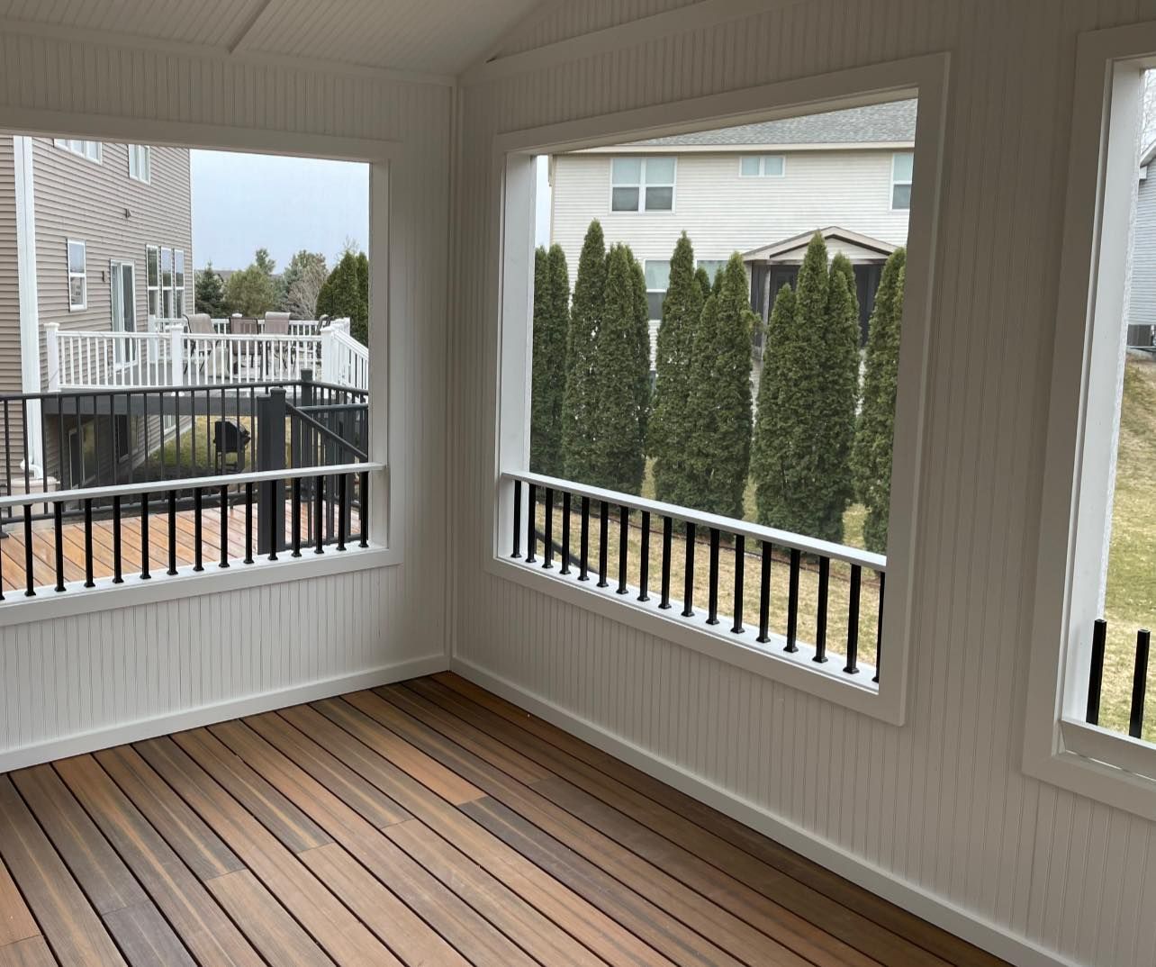 Sunroom interior with wooden floor, white walls and windows with black railing, overlooking a yard.