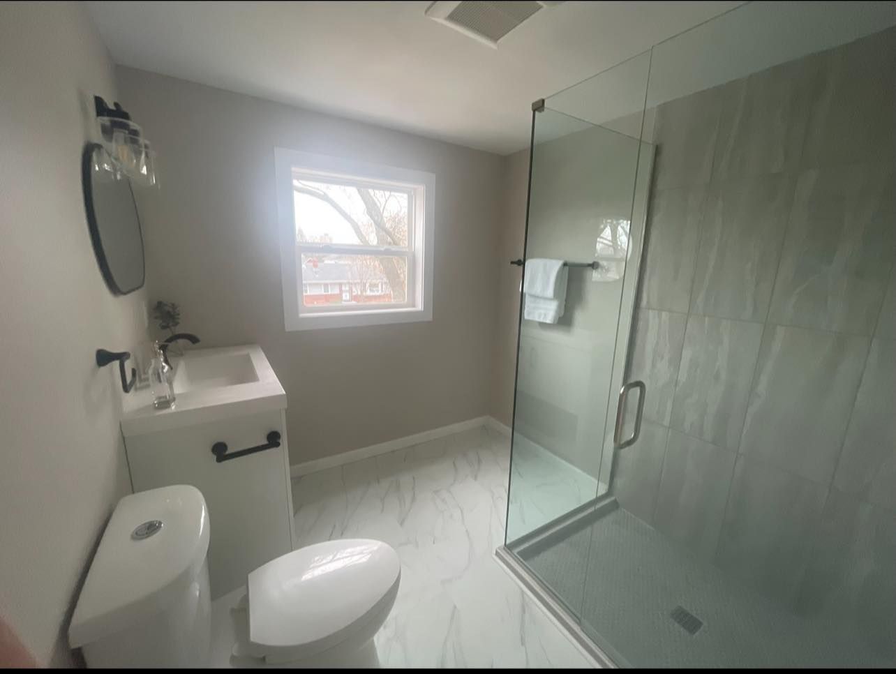 Modern bathroom with a glass shower, white vanity, and toilet, gray walls, and a window.