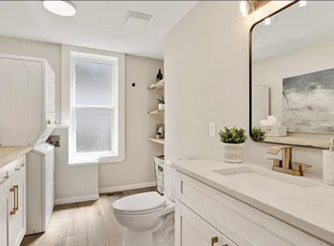 Bright white bathroom with a toilet, vanity, window, and laundry machines.
