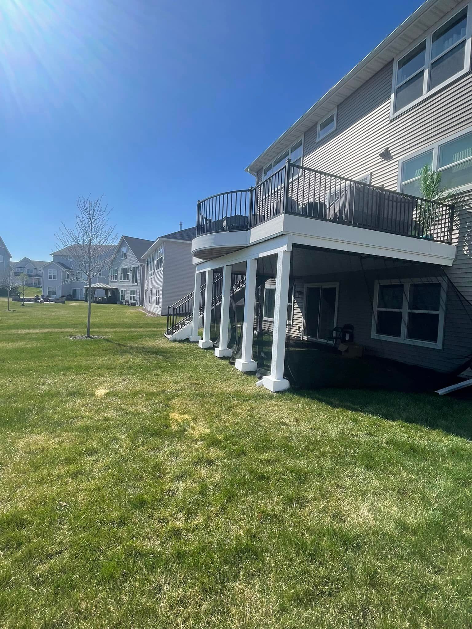 Exterior view of a two-story house with a raised deck supported by white columns, on a grassy lawn under a blue sky.