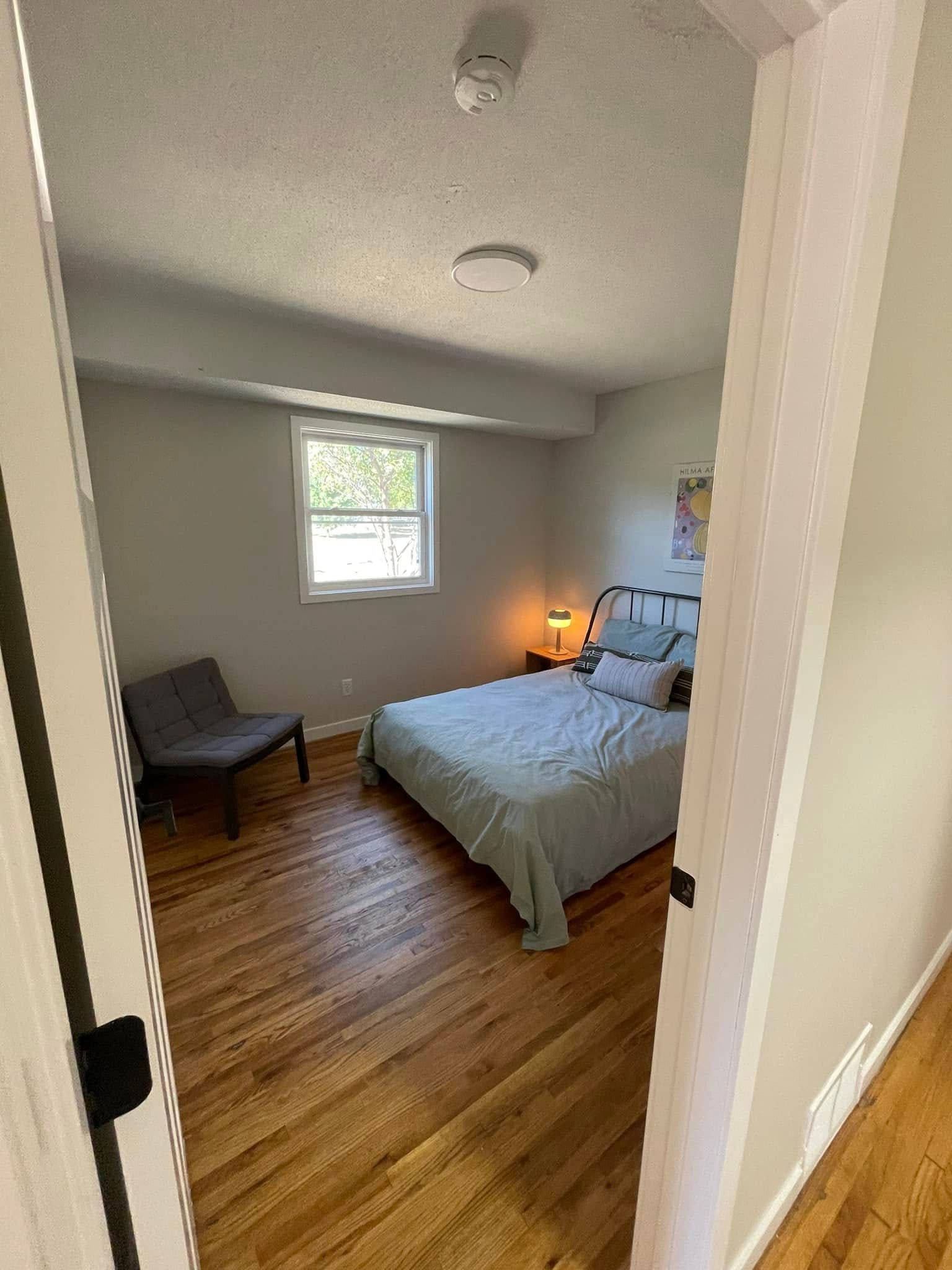 Bedroom interior with bed, chair, and window; light blue bedding, wood floors, and neutral walls.