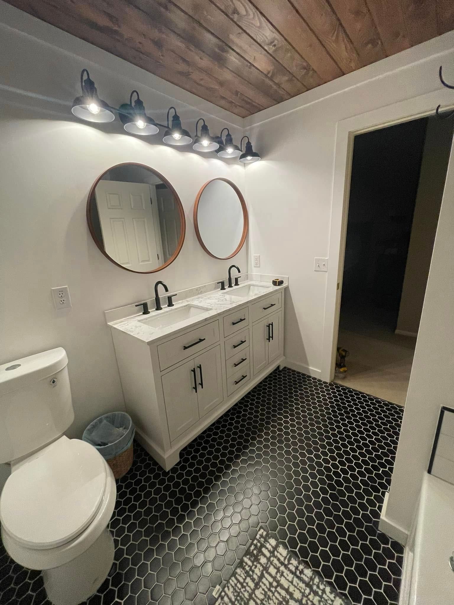Bathroom with white double vanity, round mirrors, black patterned floor, and wooden ceiling.