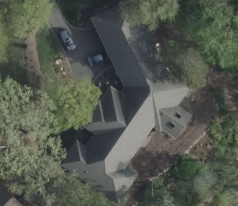 Aerial view of a dark-roofed house with a long driveway and two parked cars, surrounded by trees.