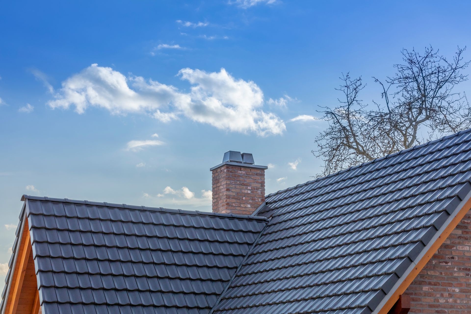 Gray tiled roof with a brick chimney against a blue sky with clouds.