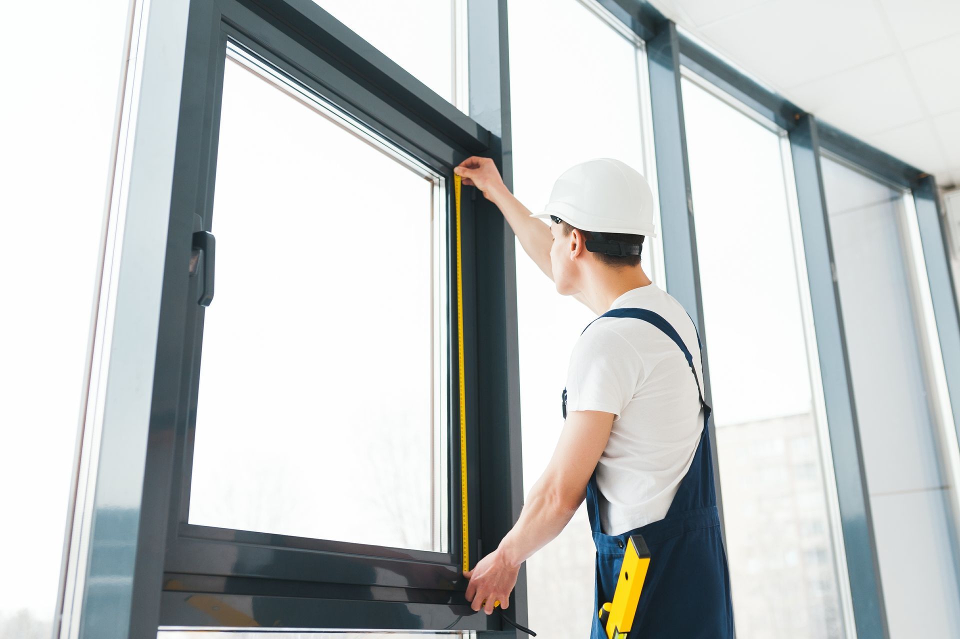 Person in hardhat measuring a window frame with a yellow tape measure inside a building.