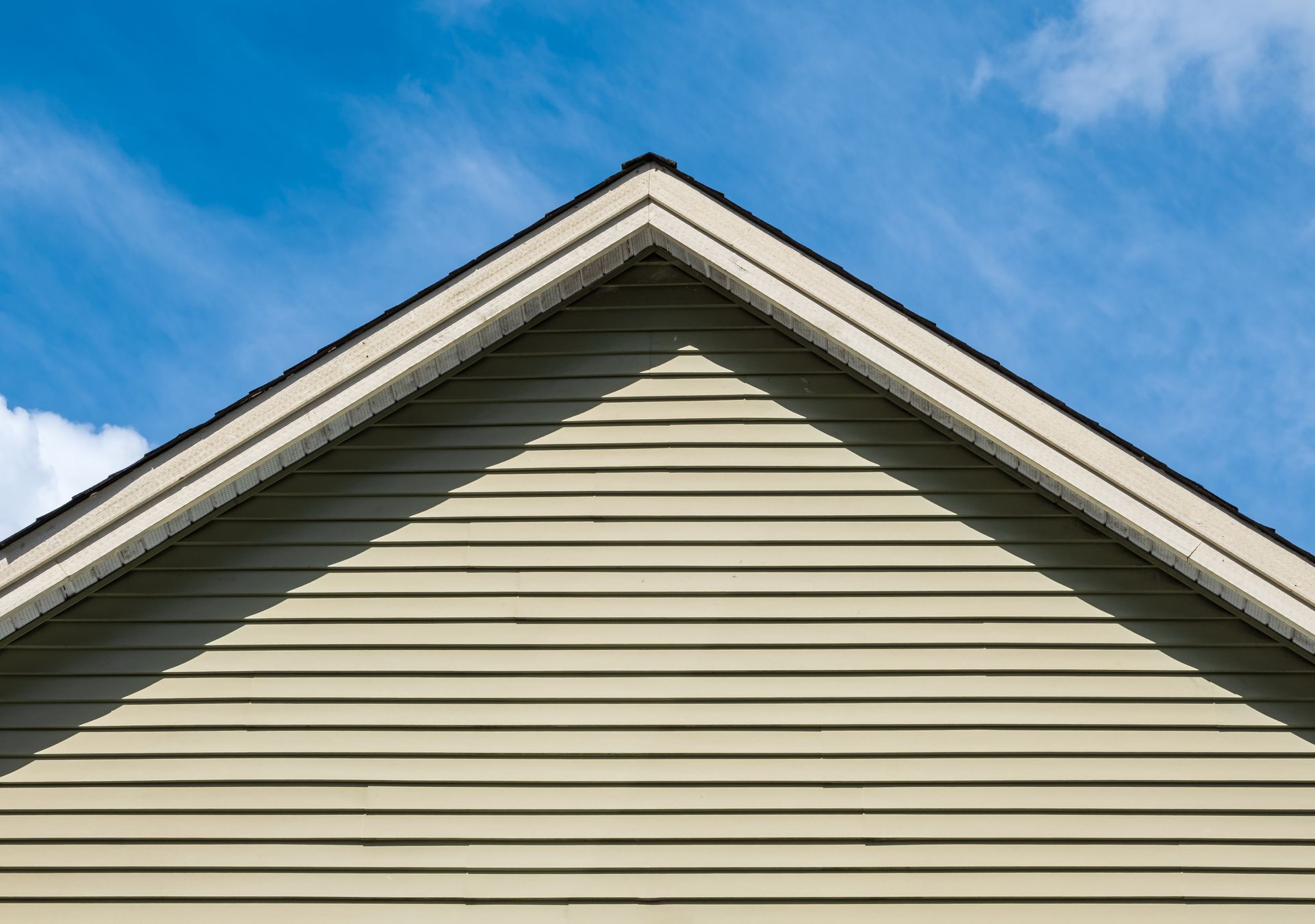 Triangular gable of a house with light tan siding against a blue sky with wispy clouds.