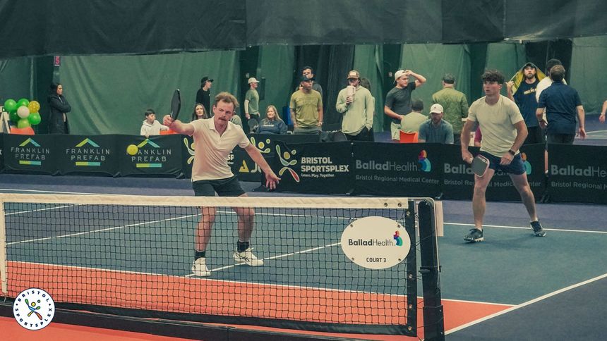 Players volley on an indoor pickleball court with spectators behind the net.