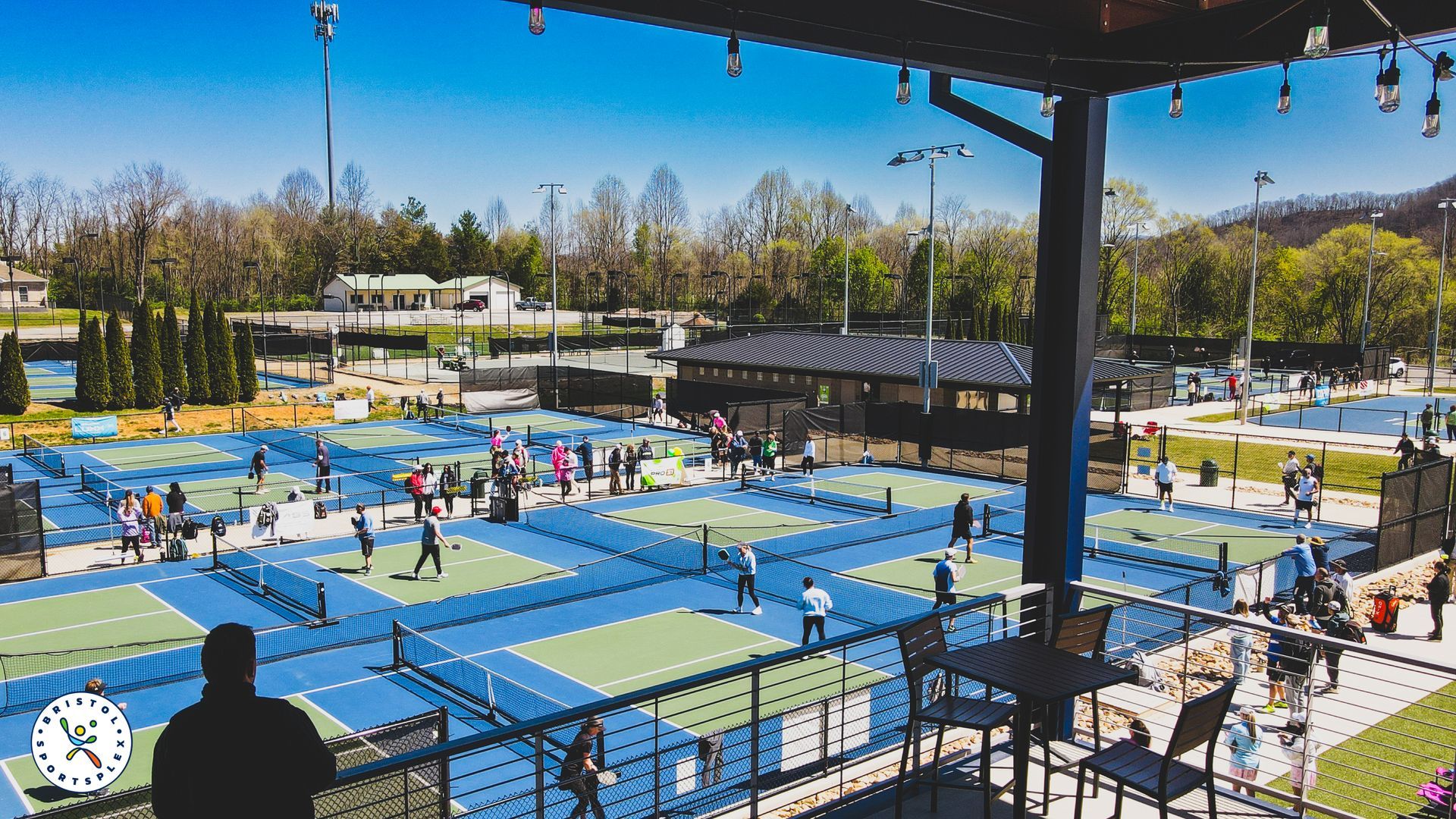 Outdoor pickleball courts with many players and spectators under a sunny blue sky viewed from VIP Section. 