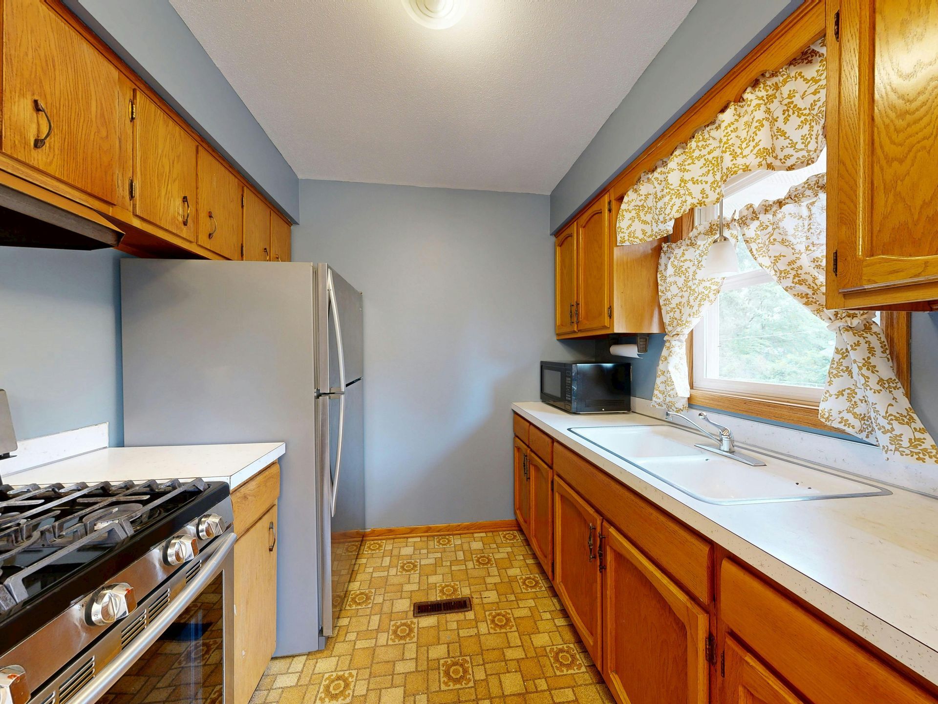 Kitchen with wooden cabinets, a stove, a refrigerator, and a window with floral curtains.