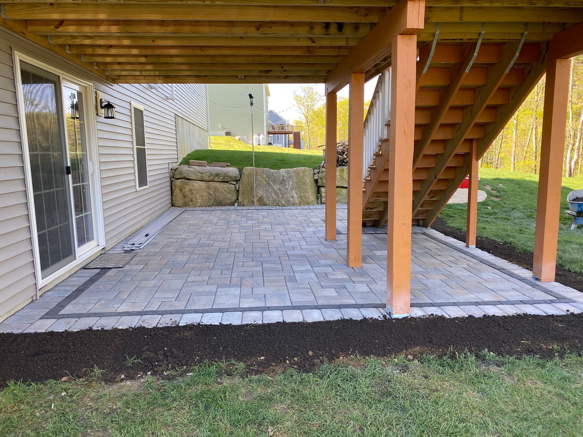 A patio under a wooden deck with stairs leading up to it.