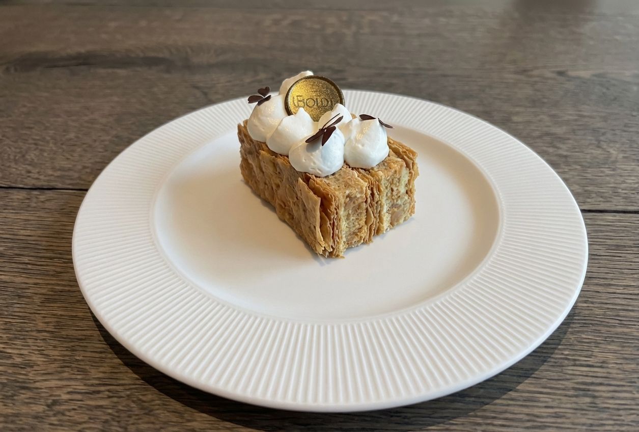 En rektangulær wienerbrød med lag af sprød dej toppet med sprøjtet fløde og en dekorativ guldmønt på en hvid tallerken.