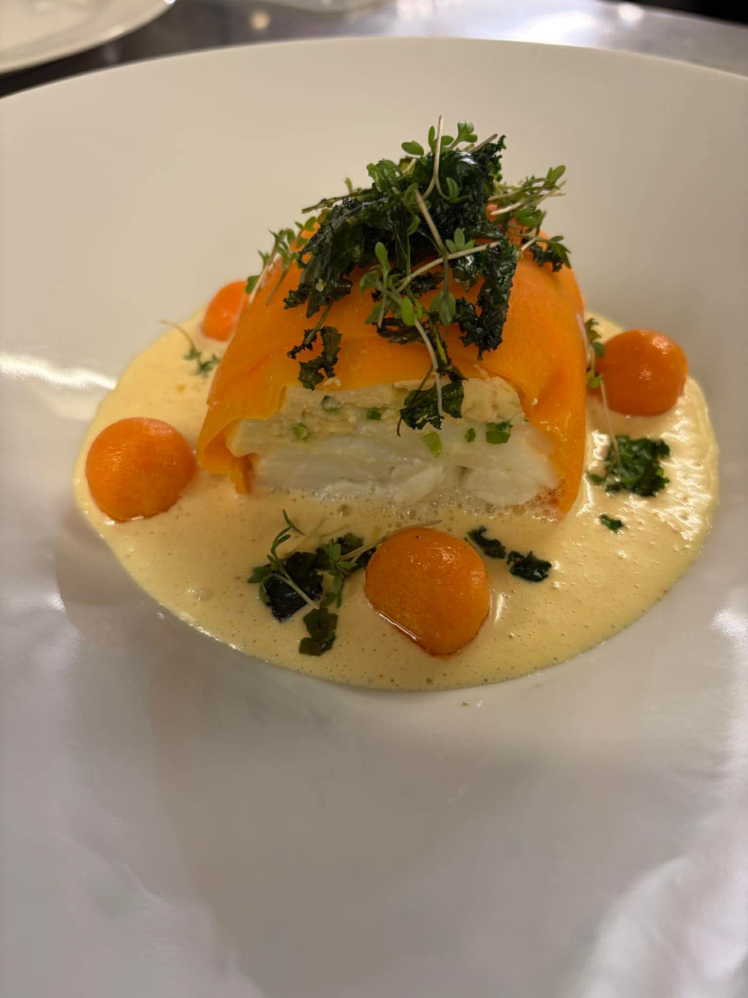 En portion hvid fisk toppet med appelsincitrusskiver, mikrogrønt og appelsinkugler i en cremet, lys gul sauce.