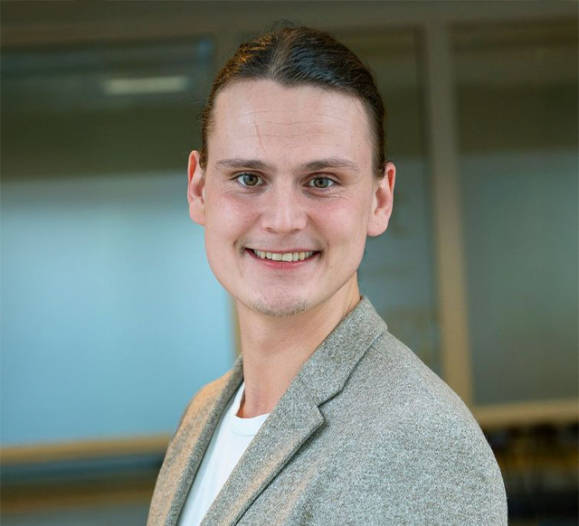 Smilende person i en grå blazer mod en sløret kontorbaggrund