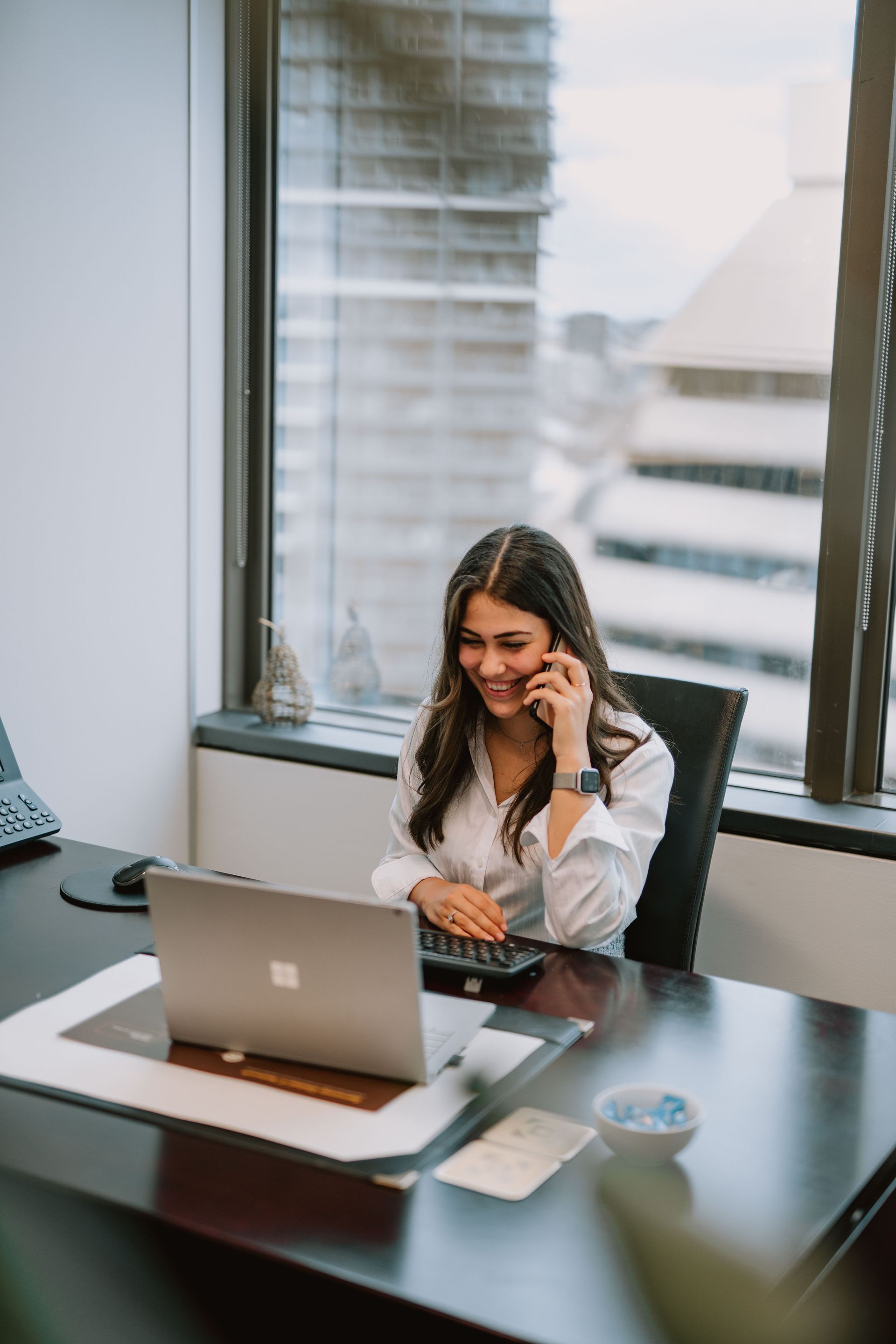 A woman is sitting at a desk with a laptop and talking on a cell phone.