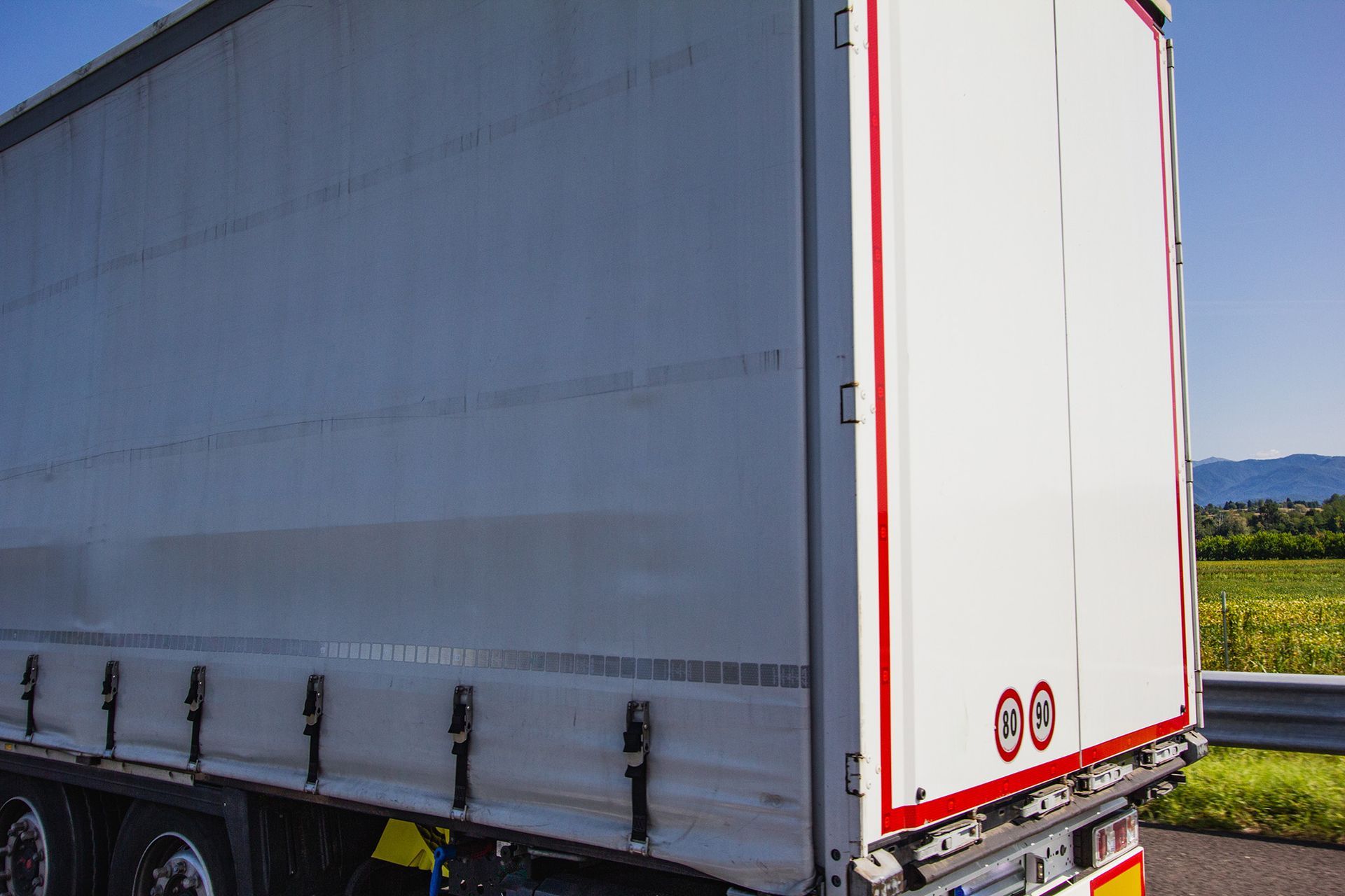 White semi-truck trailer with red trim on a road, with a field and mountains in the distance.